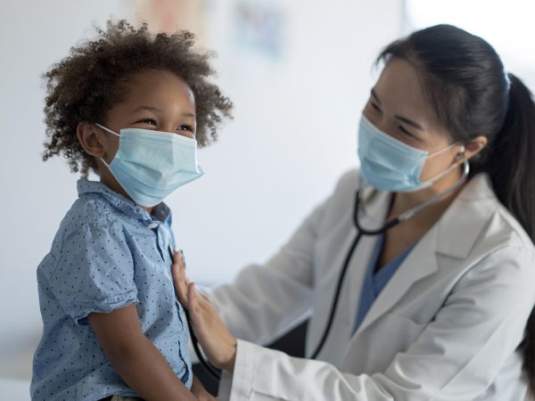 A female doctor of Asian decent holds her stethoscope to a little boys chest as she listens to his heart. The little boy is dressed casually and smiling as he sits still for the appointment.  Both the patient and the doctor are wearing medical masks to protect them from COVID.